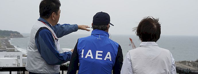 IAEA Director General and two other people stand on a roof of the Fukushima Daiichi nuclear power plant and look out over the ocean.