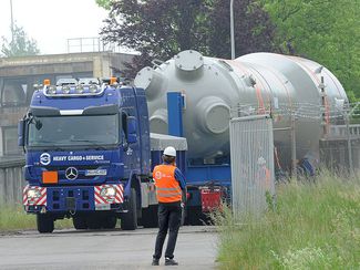 Ein sehr großes, hellgraues und röhrenartiges Gebilde aus Stahl wird auf einem blauen Schwerlast-LKW durch ein Werkstor transportiert. Davor ein Mann mit oranger Schutzweste.